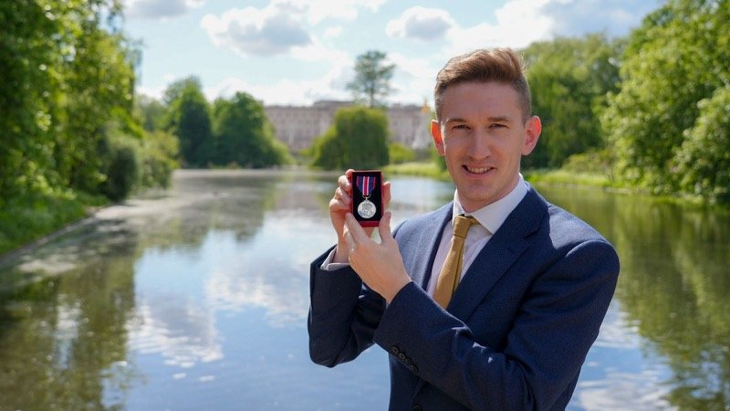 Robert Midgley holding his Coronation Medal in its case beside a lake